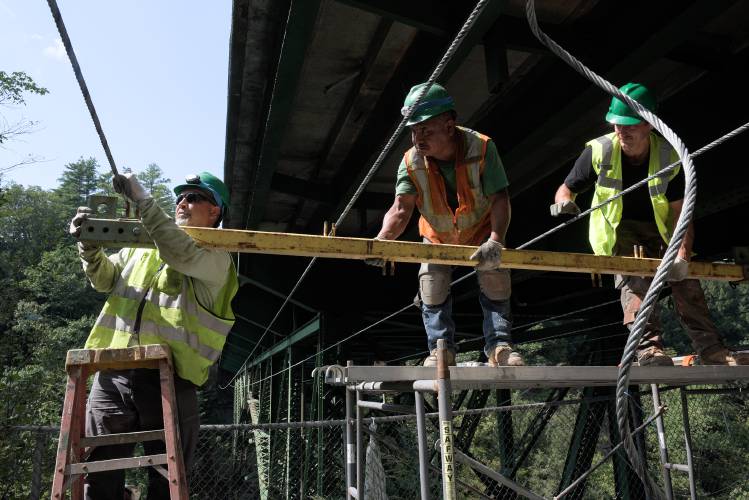 Juan Perez, left, Julio Gonzalez, middle, and James Pederson, right, of Tri-State Painting assemble cables and bracing for a containment system that will allow them to work under the Quechee Gorge Bridge and remove paint without material escaping into the river below or surrounding areas in Quechee, Vt., on Tuesday, Sept. 5, 2023. The Quechee Gorge Trail will be detoured to the Visitor Center Trail for the duration of the work. In the first phase of the project this fall, paint will be removed so the steel structure can be assessed. Over the next two years, corroded parts of the structure will be replaced, it will be repainted, the deck will be widened to accommodate six foot sidewalks that will extend west on Route 4 to parking areas, and new safety fencing will be installed, said Tom Chase, of Greenman-Pederson Inc., a consultant to the Vermont Agency of Transportation. From April to November of 2024 and 2025, automobile and pedestrian traffic will be reduced to one alternating lane. (Valley News - James M. Patterson) Copyright Valley News. May not be reprinted or used online without permission. Send requests to permission@vnews.com.