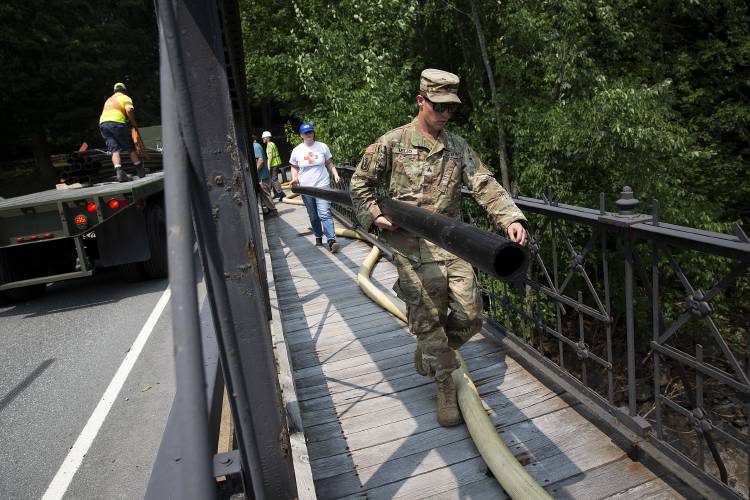 Army National Guard Sgt. Connor Mello carries a section of pipe across Elm Street Bridge with the help of Tess Malloy, who works in customer relations at the Woodstock Aqueduct Company, in Woodstock, Vt., on Monday, July 17, 2023. The potable water pipe will replace the fire hose connecting two fire hydrants across the river, allowing the town to access drinking water while the Woodstock Aqueduct Company works on a permanent solution to a broken pipe. (Valley News / Report For America - Alex Driehaus) Copyright Valley News. May not be reprinted or used online without permission. Send requests to permission@vnews.com.
