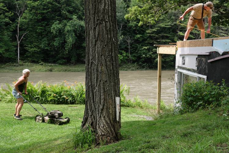  “Living on the river is worth it in retirement,” said Chris Carroll, right, who is building a home for himself and his wife Patty, left,  in the flood plain on the White River in Sharon, Vt., on Wedensday, July, 12, 2023. A stake mounted on a tree marks the high water mark from Tropical Storm Irene in the yard. “This is my last house,