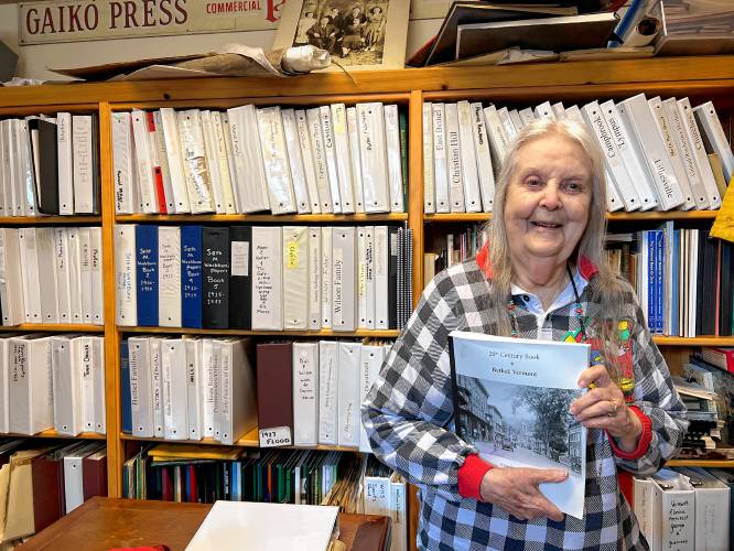 Janet Hayward Burnham stands with a copy of her book that details the history of Bethel in the 20th century. Burnham spent hours using the Bethel Historical Society's archives, organized partly in binders on the shelves behind her, to do her research. (Valley News — Liz Sauchelli)