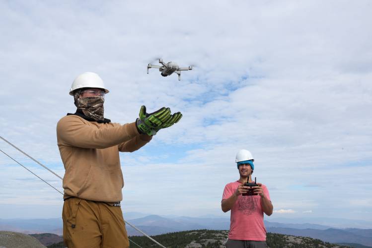 Javier Arias lands his drone in the hands of Valley Restoration co-worker Mike Morrison, left, after taking aerial video of the fire tower cab on Mount Cardigan in Orange, N.H., on Friday, Oct. 27, 2023. After more than three months of contending with weather delays, the new cab structure is in place and finish work can begin. (Valley News - James M. Patterson) Copyright Valley News. May not be reprinted or used online without permission. Send requests to permission@vnews.com.